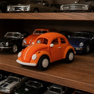 Peut inclure: Une voiture jouet orange crochetée avec des détails blancs, notamment une bande centrale et des roues à jantes blanches. La voiture est exposée sur une étagère en bois avec d'autres modèles réduits.