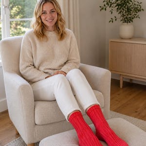 May include: A woman seated in a beige armchair wearing a cream-colored sweater, white pants, and bright red ribbed knit socks. The socks are the focal point, providing a pop of color against the neutral tones of the room and clothing. The room has a window and a side table.