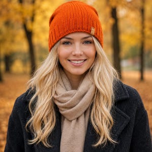 May include: A woman wearing an orange knit beanie, a beige scarf, and a dark coat. The beanie has a small brown tag. The background features blurred autumn foliage.