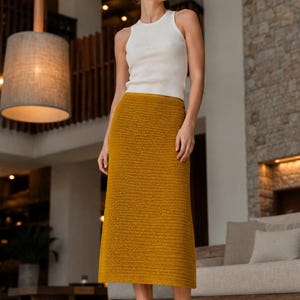 Op de afbeelding: Een wit geribbeld mouwloos hemdje gecombineerd met een okergele gehaakte midirok. De rok heeft een consistent horizontaal steekpatroon en een recht silhouet dat tot onder de knieën valt, aangevuld met eenvoudige beige platte schoenen.