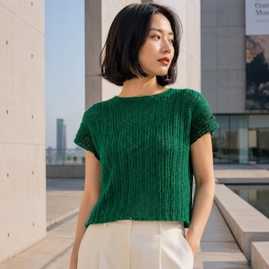 May include: A short-sleeved, emerald green knit top with a textured, open-stitch pattern and a round neckline, paired with solid white trousers. The garment features a straight hemline and a lightweight construction, providing a clean, simple look.