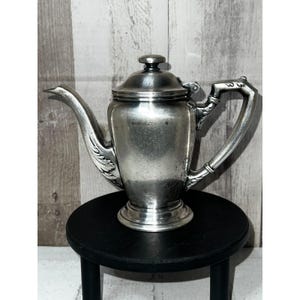 May include: A silver-toned metal teapot featuring a curved spout with embossed leaf detailing, a hinged lid with a small knob, and an angular handle. The body of the teapot has a smooth, polished finish with a pedestal base and a tapered neck.