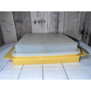 May include: A square yellow plastic storage container with a flat, translucent white lid. The container features a wide, shallow base with integrated side handles and a secure-fitting lid that sits flush within the rim of the yellow base.