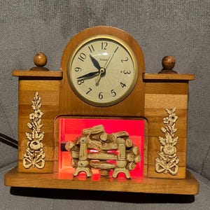 Op de afbeelding: Vintage houten mantelklok met een wijzerplaat en een decoratief haardontwerp. De klok heeft een oplichtende haardfunctie met een rode gloed. De klok is gemaakt van hout en heeft bloemaccenten aan de zijkanten.