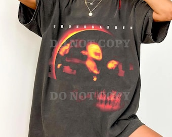 May include: A charcoal grey short-sleeve t-shirt featuring the text SOUNDGARDEN in white letters above a graphic of a glowing orange and red sun-like arc and a blurred figure. The shirt has a crew neckline and a loose, relaxed fit throughout.