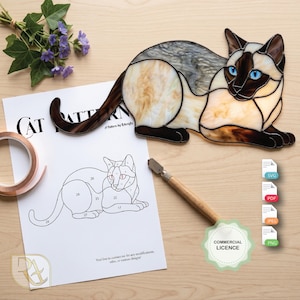 May include: A stained glass Siamese cat figure with blue eyes, cream, brown, and grey marbled glass pieces. Beside it lies a paper sheet titled CAT PATTERN with a numbered line drawing, a roll of copper foil tape, and a glass cutting tool.