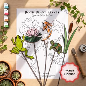 Puede incluir: Una hoja de patrón de vidrieras titulada Pond Plant Stakes con dibujos lineales de una rana, loto, pez koi, tortuga y espadaña. Cuatro piezas de vidriera terminadas se muestran sobre el patrón, incluyendo una rana verde, un loto rosa, un koi naranja y blanco, y una tortuga verde.
