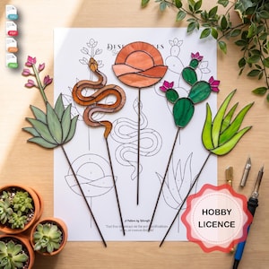 Puede incluir: Cuatro estacas de vidrio tintado con temática del desierto que incluyen una serpiente, un sol, un cactus y una planta de aloe, sobre una hoja de patrón impresa. Una etiqueta roja festoneada con el texto HOBBY LICENCE es visible.