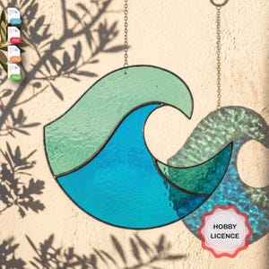 May include: A stained glass suncatcher shaped like a stylized ocean wave, featuring segments of light green, bright blue, and dark teal glass held together by dark metal solder lines, suspended by a thin metal chain against a textured wall.