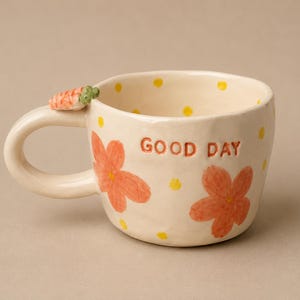 May include: Cream-colored ceramic mug with orange floral designs and yellow polka dots. The mug has the words "GOOD DAY" and a carrot-shaped handle accent. A charming and cheerful design for a coffee or tea cup.