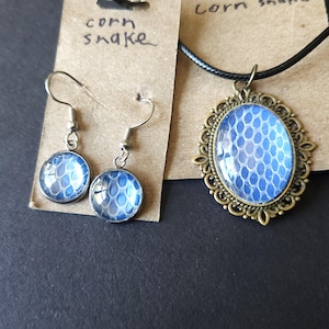 May include: A jewelry set featuring a pendant necklace and matching earrings. The pendant and earrings have a blue and white pattern, resembling snake scales, set in a bronze-colored frame. The earrings hang from silver-colored hooks.