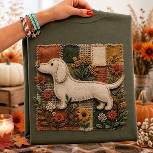 May include: An olive green sweatshirt featuring a central rectangular patch with a beige fabric dachshund appliqué. The design includes embroidered flowers in shades of orange, yellow, and white, set against a background of textured fabric squares.