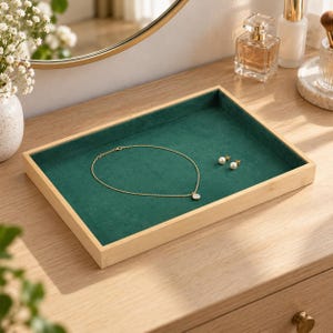 May include: A rectangular wooden jewelry tray featuring a deep green velvet lining. The tray contains a delicate gold chain necklace with a small circular pendant and a pair of white pearl stud earrings resting on the soft fabric surface.