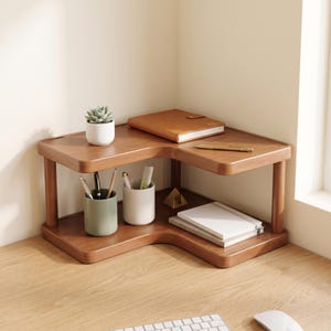 May include: A two-tiered L-shaped wooden corner desk organizer with rounded edges. The top shelf holds a small potted succulent and a tan notebook. The bottom shelf contains two cylindrical pen holders, one sage green and one white, and a stack of notebooks.