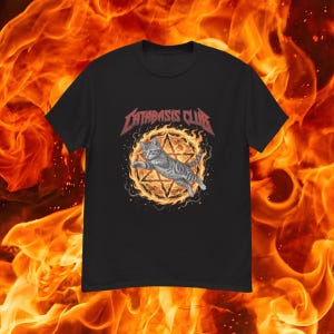 Pode incluir: Uma t-shirt preta de manga curta com uma estampa gráfica de um gato malhado cinzento a saltar através de uma estrela em chamas. Acima do gato, o texto CATABASIS CLUB está impresso numa fonte vermelha estilizada no peito da t-shirt.