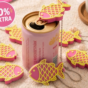 May include: A pink aluminum beverage can with the text MISCHGETRÄNK, surrounded by several pink and yellow fish-shaped silicone tab openers attached to silver ball chains. A circular pink graphic in the corner displays the text 10% EXTRA.