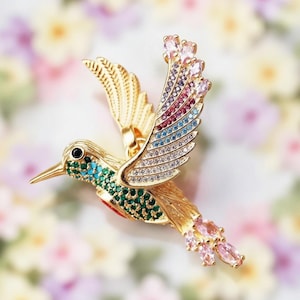 May include: A gold hummingbird brooch with colourful gemstones. The wings are decorated with a gradient of pink, purple, blue, and clear stones. The body features green and turquoise accents, and the tail has pink stones.