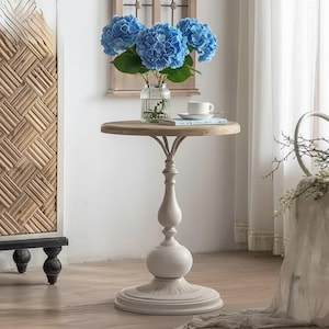 Può includere: Un tavolino rotondo con piano in legno chiaro e base bianca ornata. Il tavolo ospita un vaso di vetro con ortensie blu e una tazza da tè bianca. Il tavolo è alto circa 61 cm.