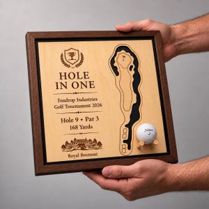 Könnte beinhalten: Eine quadratische Holzrahmentafel mit einer hellen Holzplatte, einer lasergravierten Golfplatzkarte, einem weißen Titleist-Golfball auf einem kleinen Holz-Tee sowie dem Text HOLE IN ONE, Foxdrop Industries Golf Tournament 2026, Hole 9, Par 3, 168 Yards und Royal Bromont.