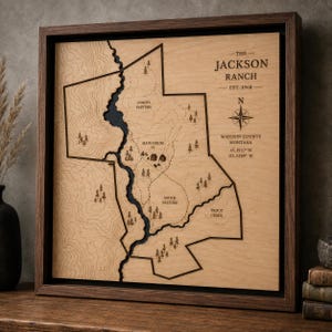 May include: A square wooden map of The Jackson Ranch, Est. 1968, in Madison County, Montana. The light wood surface features engraved topography, tree icons, property boundaries, and labels for North Pasture, Main House, South Pasture, and Trout Creek.