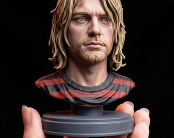 Könnte beinhalten: Eine detaillierte Büste einer Person mit schulterlangem blonden Haar und leichtem Bart, bekleidet mit einem Shirt mit horizontalen roten und schwarzen Streifen. Die Figur steht auf einem soliden, runden grauen Sockel mit realistischen Zügen.