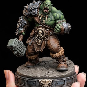 Könnte beinhalten: Detaillierte Figur eines grünschlägigen Ork-Kriegers mit bedrohlichem Gesichtsausdruck. Der Ork ist in brauner Leder- und Metallrüstung gekleidet und hält einen großen Hammer. Die Basis ist eine steinartige Plattform mit dekorativem Rand.