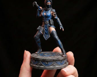 May include: A detailed figurine of a female character in a blue and black outfit, holding a curved blade. The figure stands on a circular, ornate base with a blue design. The character is wearing a mask and has a determined expression.
