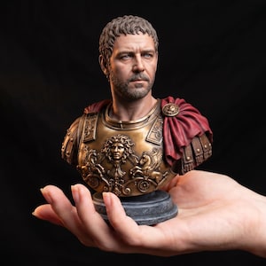 May include: A detailed bust of a man with short brown hair and a beard, wearing bronze-coloured armour with a central Gorgon emblem and a draped red cloak over the left shoulder, set upon a circular dark grey base against a black background.