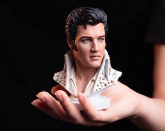 May include: A detailed bust of Elvis Presley featuring his signature black pompadour hairstyle, blue eyes, and a high-collared white jumpsuit adorned with gold star-shaped studs. The sculpture is rendered in a realistic style with a smooth finish.