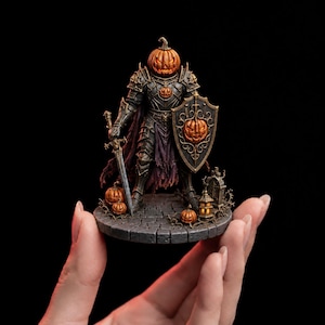May include: A miniature figurine of a warrior with a carved pumpkin head, wearing dark ornate armor and a tattered purple cape. The figure holds a sword and a shield featuring a pumpkin emblem, standing on a stone base with small pumpkins and a tombstone.