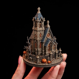 Pode incluir: Um modelo de igreja gótica em miniatura com paredes de textura de pedra, um telhado de telhas azul-escuras e uma torre central alta. A estrutura é cercada por uma cerca de metal preta, uma árvore nua e pequenas abóboras laranja na base.