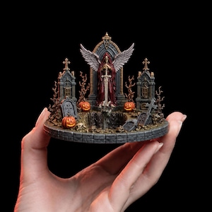 Może przedstawiać: Miniaturowa diorama przedstawiająca centralną postać w kapturze z dużymi skrzydłami trzymającą miecz, umieszczoną przed gotyckim łukiem. Scena zawiera dwa kamienne mauzolea, kilka nagrobków, bezlistne drzewa i trzy pomarańczowe dynie.