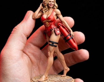 May include: A figurine of a blonde woman in a red Baywatch swimsuit, holding a red rescue buoy. The figure has a detailed face and is standing on a textured base. The swimsuit has the Baywatch logo.