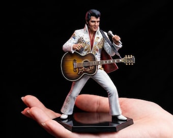 May include: A figurine of a performer with dark hair, wearing a white jumpsuit with gold and red accents, holding a silver microphone in one hand and playing a brown acoustic guitar. The figure stands on a black hexagonal base against a black background.