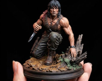 May include: A detailed figurine of a kneeling action figure, holding a knife. The figure is wearing a brown tank top, cargo pants, and boots. A red headband is visible. The figure is posed on a base with a tree trunk and foliage.