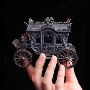 Può includere: Un modello di carrozza in miniatura grigio scuro con intricati motivi a volute, finestre ad arco e decorazioni a forma di pipistrello sul tetto. La carrozza presenta quattro ruote a raggi, un sedile per il conducente e dettagliati rilievi.