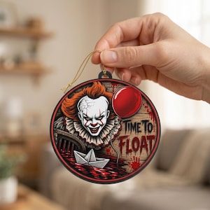 May include: A round ornament featuring a graphic illustration of a clown with orange hair, white face paint, and a ruffled collar. The design includes a red balloon, a paper boat in red liquid, and the text "TIME TO FLOAT" in a distressed font.