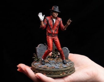 May include: A detailed figurine of Michael Jackson wearing a red leather jacket with black stripes, black pants, a white glove, and a black fedora. The figure stands on a circular base featuring two gray tombstones with the text Michael Jackson Thriller.
