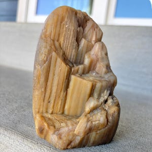 May include: A vertical, amber-colored mineral specimen with a translucent, crystalline structure. The stone features prominent, parallel vertical striations and jagged, layered formations that create a textured, multi-faceted surface appearance.