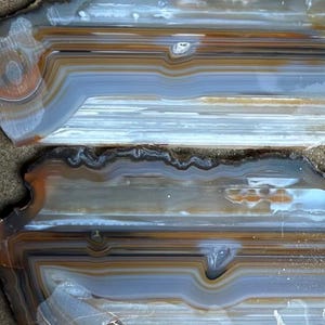 May include: Two polished agate slices featuring horizontal bands of translucent grey, white, and earthy brown tones. The stones exhibit natural mineral layering with smooth, glossy surfaces and irregular, jagged edges along the outer perimeters.
