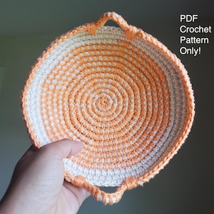 May include: A round, hand-crocheted basket featuring a spiral pattern of alternating white and orange yarn. The basket has two small, integrated handles on opposite sides. The text "PDF Crochet Pattern Only!" is printed in white on the right side.