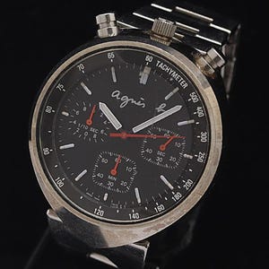Peut inclure: Montre chronographe à cadran noir avec boîtier et bracelet argentés. Le cadran comporte plusieurs sous-cadrans, des aiguilles blanches et rouges, et la marque "agnes b.". Elle comprend une échelle tachymétrique. Fond sombre.
