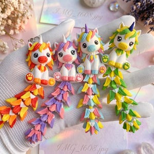 Op de afbeelding: Vier handgemaakte eenhoornbedels van polymeerklei met witte lichamen, kleurrijke manen en lange, beweegbare staarten. Elke eenhoorn heeft een uniek kleurenschema: oranje en rood, paars en roze, pastel regenboog, en groen en geel.