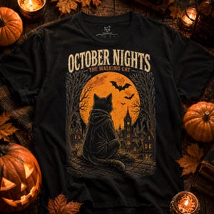 Puede incluir: Una camiseta negra con un estampado gráfico de un gato con sudadera sentado en un tejado, mirando una gran luna naranja, murciélagos y una ciudad gótica. El texto dice OCTOBER NIGHTS y THE WALKING CAT en tonos naranja, amarillo y negro.