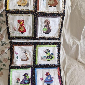 Puede incluir: Dos tapices rectangulares de tela con una cuadrícula de cuatro cuadrados blancos cada uno. Cada cuadrado contiene una figura bordada con capota y vestido, rodeada de varios motivos como flores, lluvia, un flamenco y un corazón.