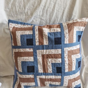 Puede incluir: Un cojín cuadrado con un diseño de patchwork tipo cabaña de troncos. El patrón consiste en tiras alternas de tela azul sólido, marrón sólido y una tela color crema con un pequeño estampado floral, acentuado por pequeños cuadrados de tela azul oscuro con puntos blancos.