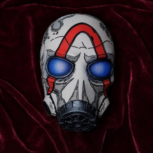 May include: A full-face mask featuring a white base with black comic-style outlines, prominent red markings across the forehead and cheeks, and two large, vibrant blue mesh eye lenses. A black circular filter component is attached at the mouth area.