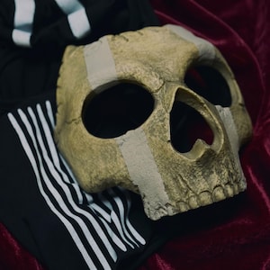May include: A gold-colored skull mask with large eye sockets and a prominent nose cavity. The mask has a weathered appearance and is secured with tan tape. Black athletic pants with white stripes are visible in the background. The mask rests on a deep red velvet surface.