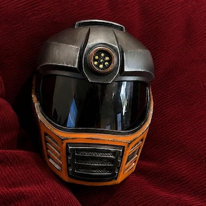 Pode incluir: Um capacete integral com uma secção superior em prata metálica com uma luz circular, uma viseira escura e uma secção inferior em laranja brilhante com grelhas de ventilação pretas. O capacete apresenta um acabamento desgastado e escuro.
