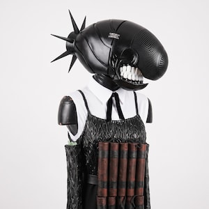 Puede incluir: Un casco negro con púas, frente perforada y dientes blancos, sobre una camisa blanca de cuello con corbata negra y un vestido negro texturizado que presenta una fila de objetos cilíndricos marrones similares a cartuchos de dinamita.